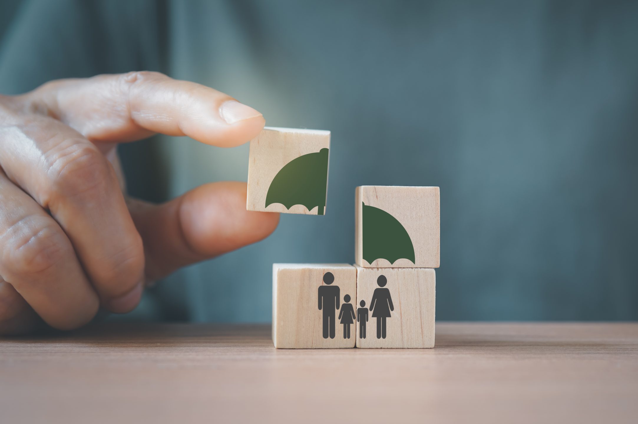 Senior's Hand Complete Umbrella Over Family Icon On Wooden Cube Blocks, For Life Or Group Insurance